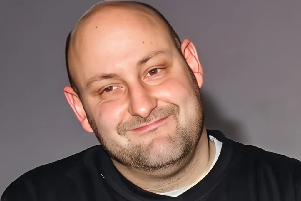 A close-up of one of the actors during rehearsal, head tilted with a wry half-smile, in a black V-neck top against a plain grey wall.