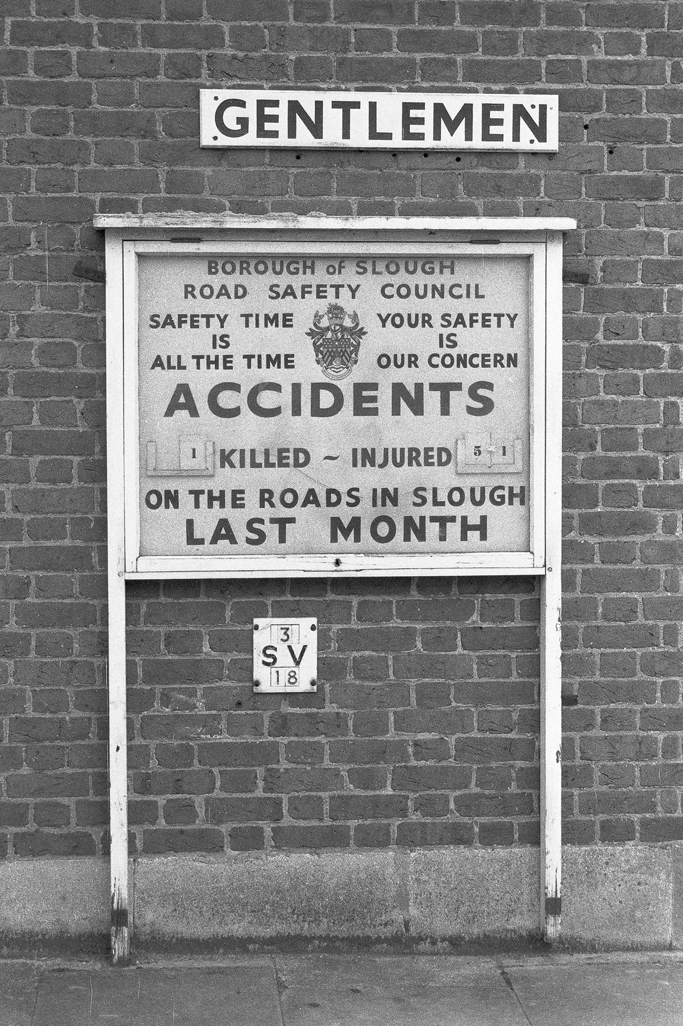 Black-and-white photograph of a brick wall with a framed roadside notice beneath a sign reading Gentlemen. The notice, issued by the Borough of Slough Road Safety Council, displays road accident statistics stating Accidents: 1 killed, 51 injured on the roads in Slough last month, with an official crest at the centre.