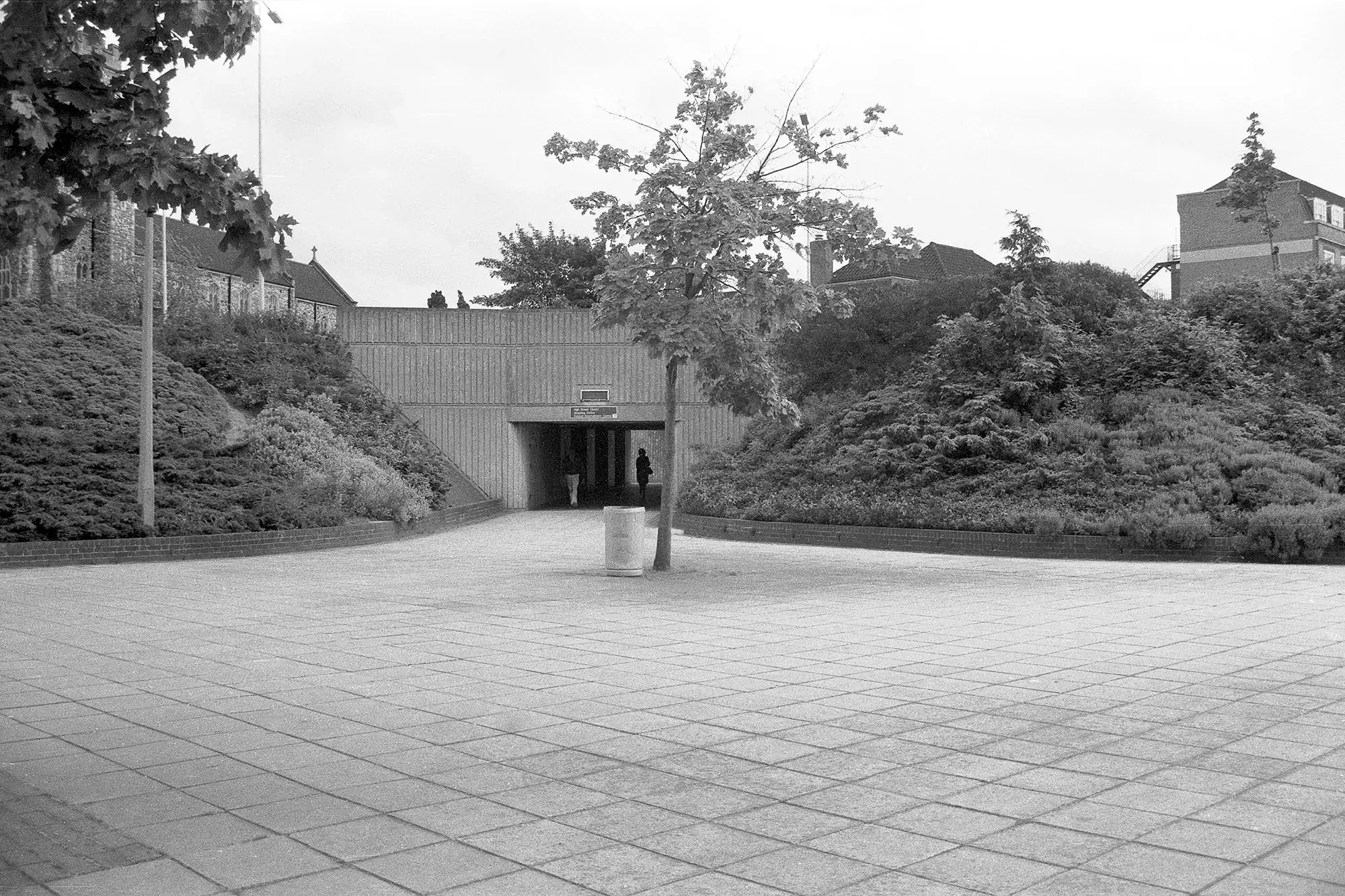 Black-and-white photograph inside Slough's Brunel roundabout, June 1986, showing a paved pedestrian space with a small tree in the foreground and a concrete underpass leading to the High Street East, Shopping Centre, and Fulcrum Entertainment Centre. Two figures are visible walking through the underpass, with planted embankments and buildings above.