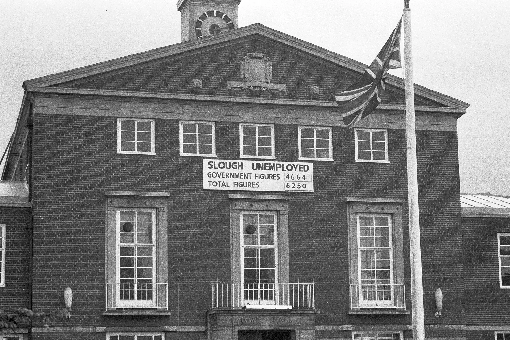 Black-and-white photograph of Slough Town Hall in 1986, showing a symmetrical brick façade with sash windows and a clock tower. A large sign titled Slough Unemployed displays government and total unemployment figures, with a Union flag flying on a pole in the foreground.