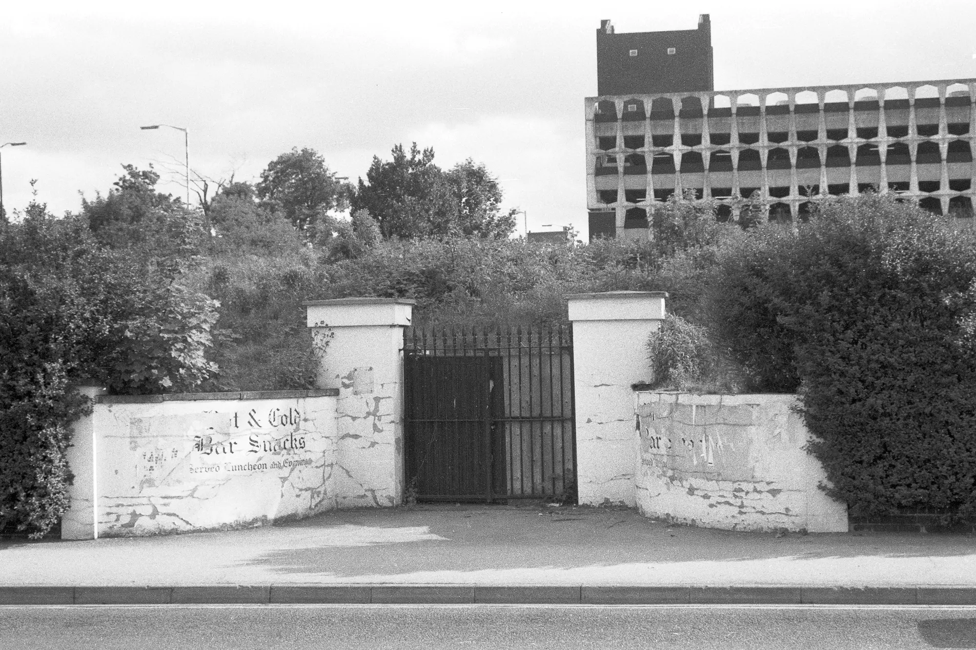 Black-and-white photograph of an overgrown gated entrance opposite Slough railway station, June 1986, with peeling painted signs on low walls, dense vegetation behind iron gates, and the Brunel Car Park and Bus Station visible in the background.