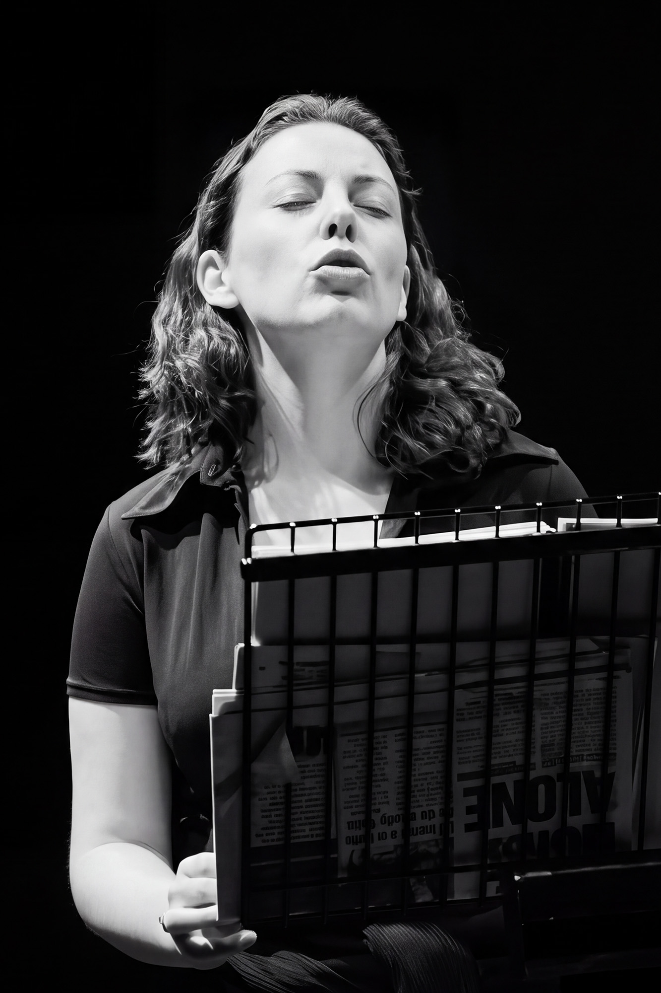 Black-and-white photograph of a person standing behind a music stand holding several printed pages. The person's head is tilted upward with eyes closed. Strong stage lighting illuminates the face, upper body, and hands, while the background remains dark. The music stand partially obscures the lower half of the body.