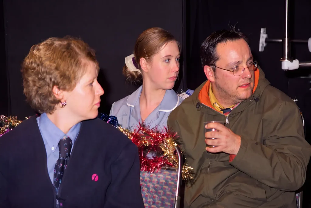 Three performers sitting on a bus seat hung with red and gold tinsel: in front a woman in a bus driver's uniform and tie, behind her a young woman in a pale uniform, and a man in glasses and a khaki parka holding a cup