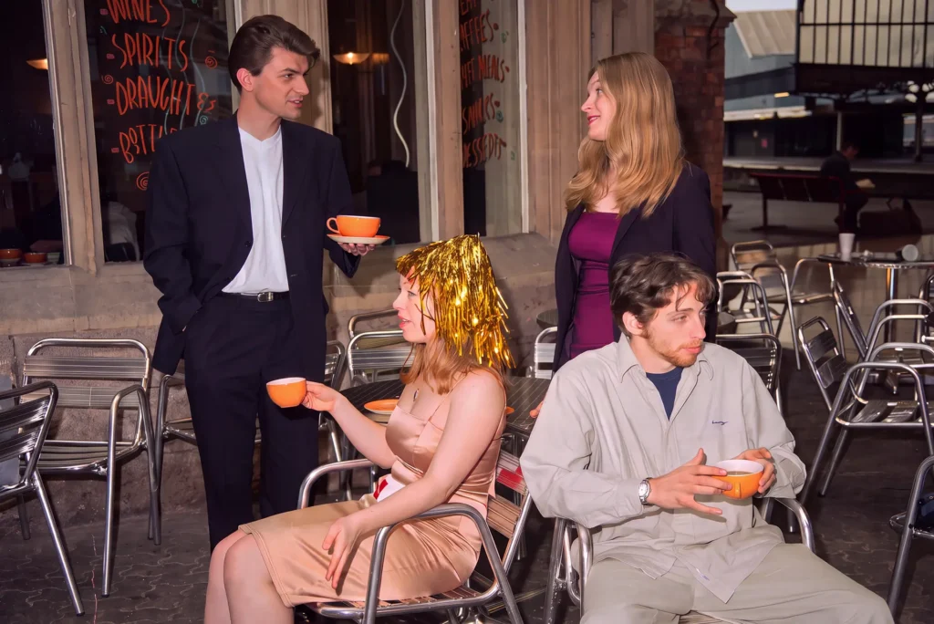 Four performers outside a bar with drinks chalked on the window, holding orange cups and saucers among metal caf&eacute; chairs; the woman seated at the front wears a gold tinsel wig