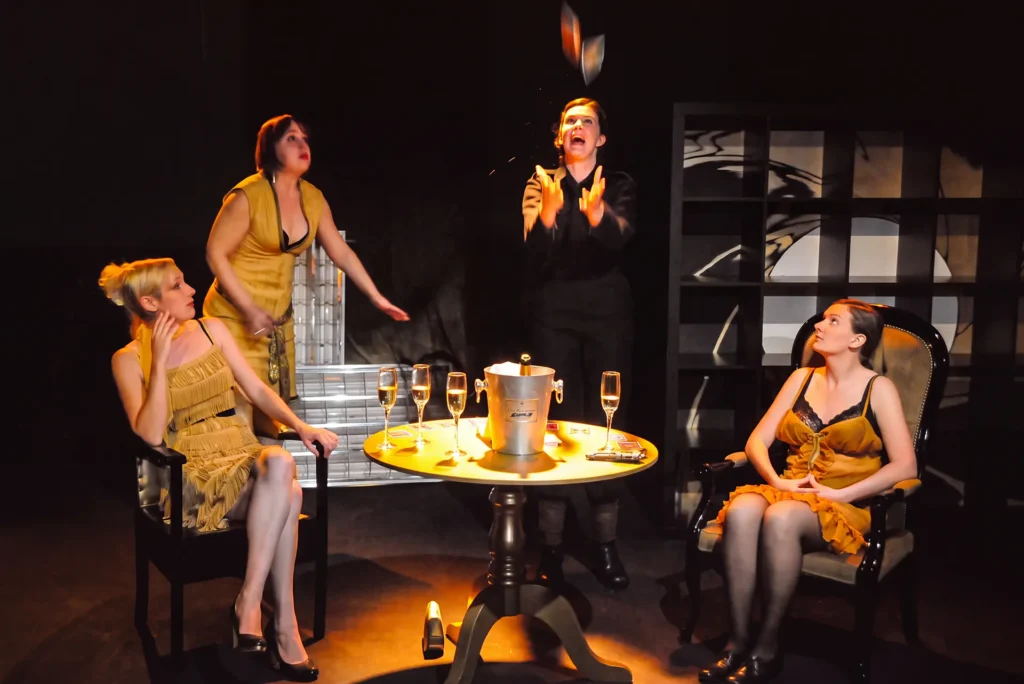 Four performers on a dark stage. Two sit in armchairs either side of a round table holding a champagne bucket, filled flutes and a golden gun; behind them a third hurries towards a fourth, who is flinging Tarot cards into the air
