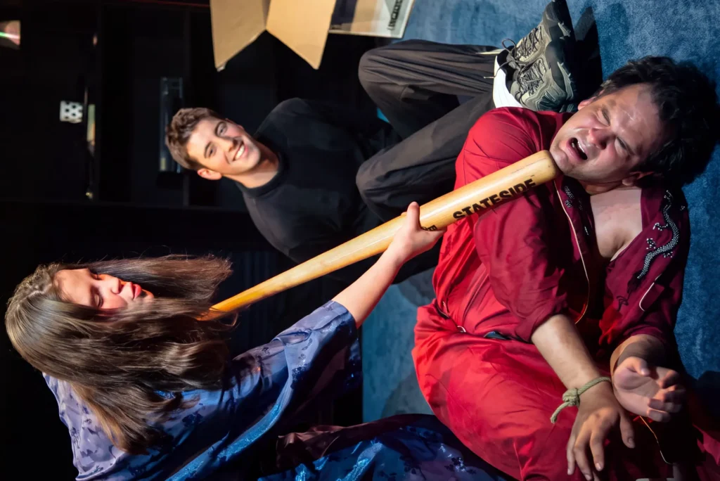 A woman in a blue dressing gown thrusts a baseball bat under the chin of a man in a red dressing gown, sprawled backwards with his wrists tied, while a third performer grins in the background