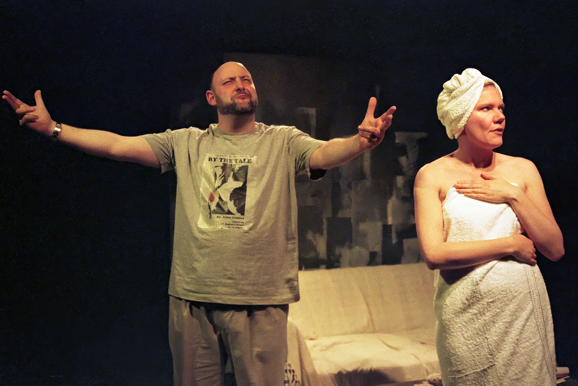 A bearded man in a grey T-shirt throws his arms wide on a dark stage; beside him a woman wrapped in a white bath towel, hair turbaned in a second towel, rests a hand on her chest