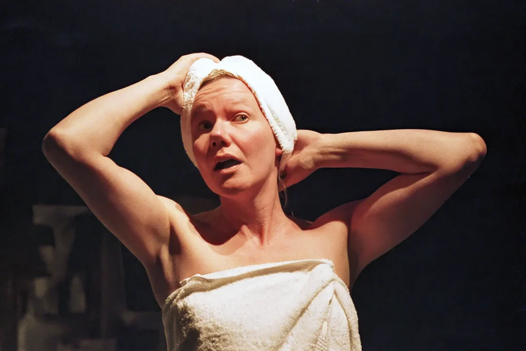 A woman on a dark stage wrapped in a white bath towel, a second towel turbaned round her head, both arms raised to it, caught open-mouthed