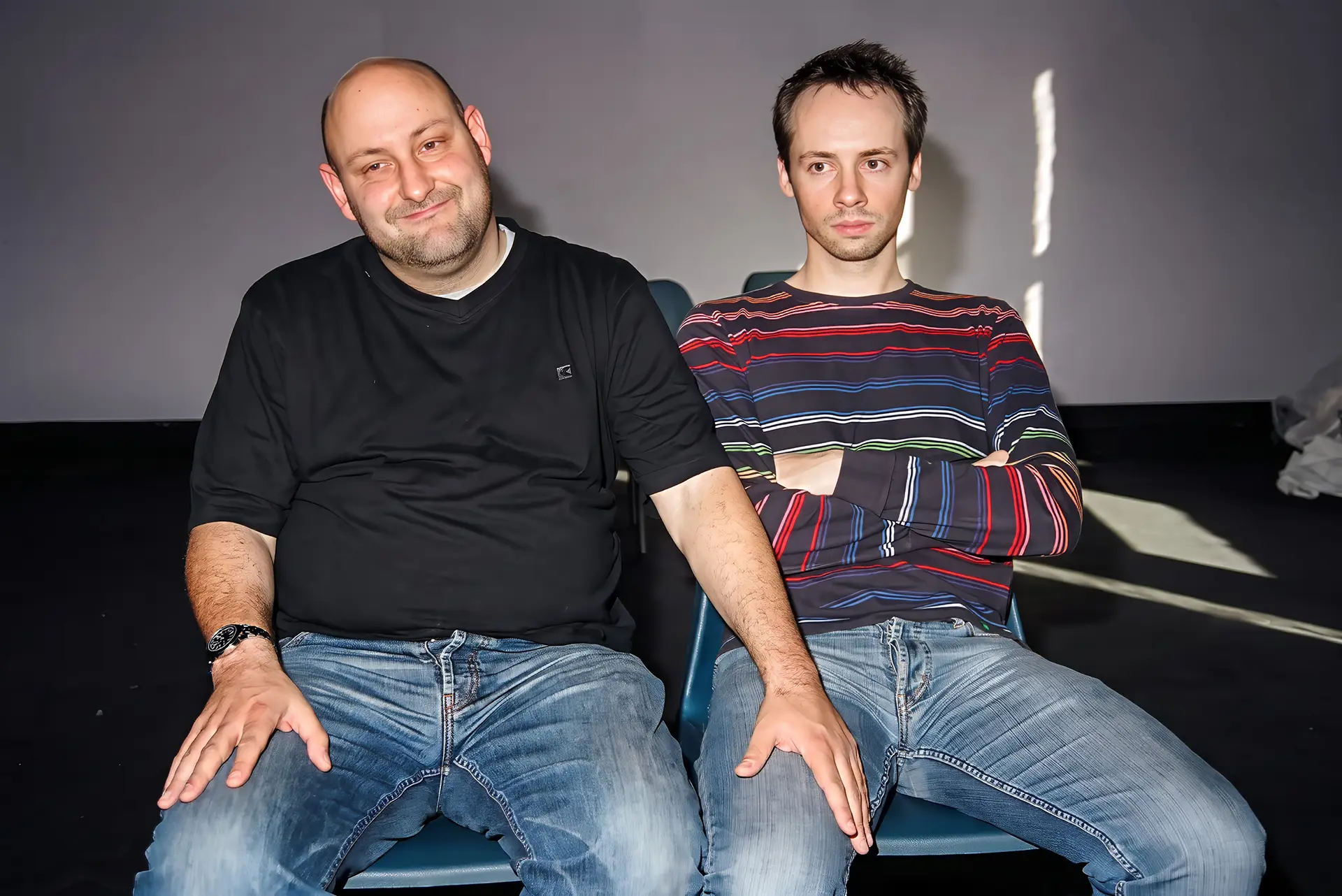 Two actors seated side by side as if in the front seats of a car, rehearsing the Arthur's Seat scene: the man on the left has a hand on the other's thigh, while the man on the right sits with his arms folded.