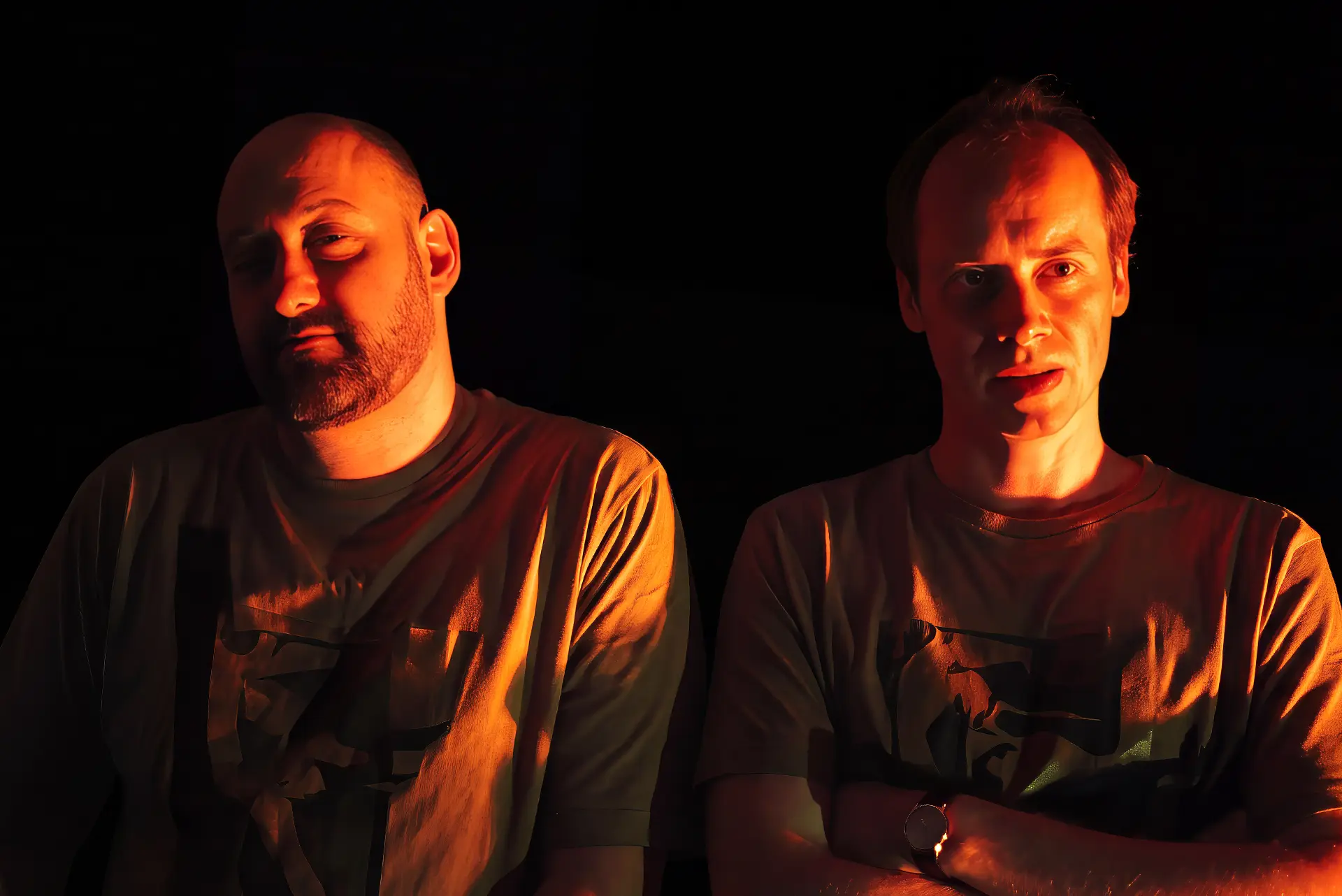 Two men side by side against a black background, lit from the right by a deep orange light that rakes across their faces and shoulders. Both wear pale printed T-shirts. The man on the left is bald with a greying beard, his eyes lowered, and his mouth closed. The man on the right has his arms folded, a round wristwatch on his wrist, and his lips parted as he looks ahead.