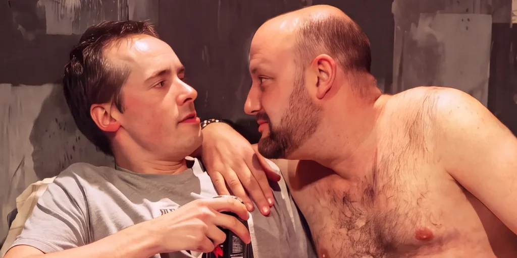 Two men close together on a pale sofa in front of grey painted flats, under warm stage light. The man on the left wears a grey t-shirt and faded blue jeans and holds a drinks can in both hands. The man on the right is bare-chested and leans in on the other man's left shoulder until their faces almost touch.