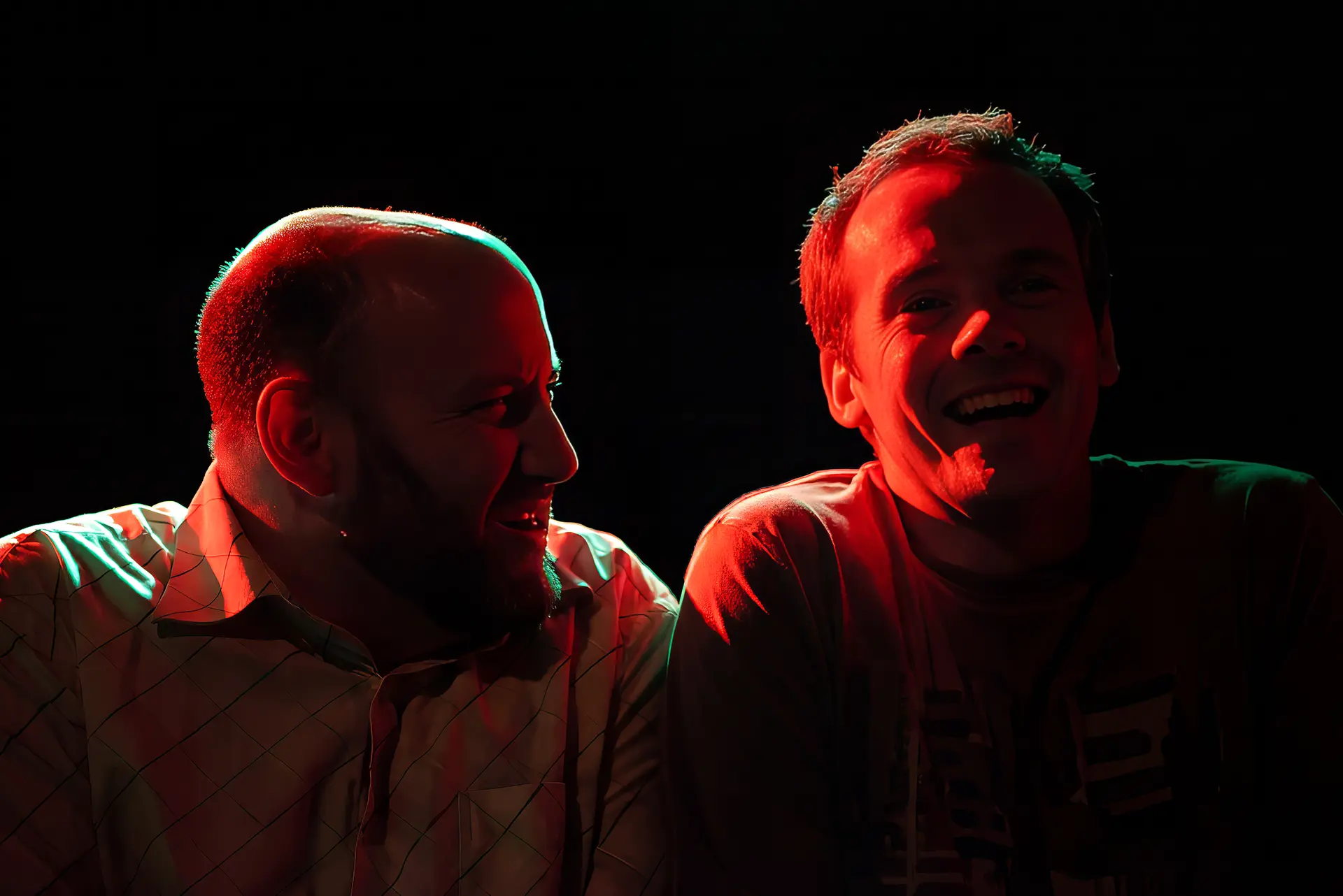 Two actors playing the ceilidh scene as a crowded bar, in costume under red stage light with a green rim along their heads and shoulders. The man on the left, in a pale checked shirt, has his head turned towards the other and tilted down, his eyes screwed shut and his teeth bared. The man on the right, in a pale t-shirt, has his head tipped towards him and his mouth wide open.