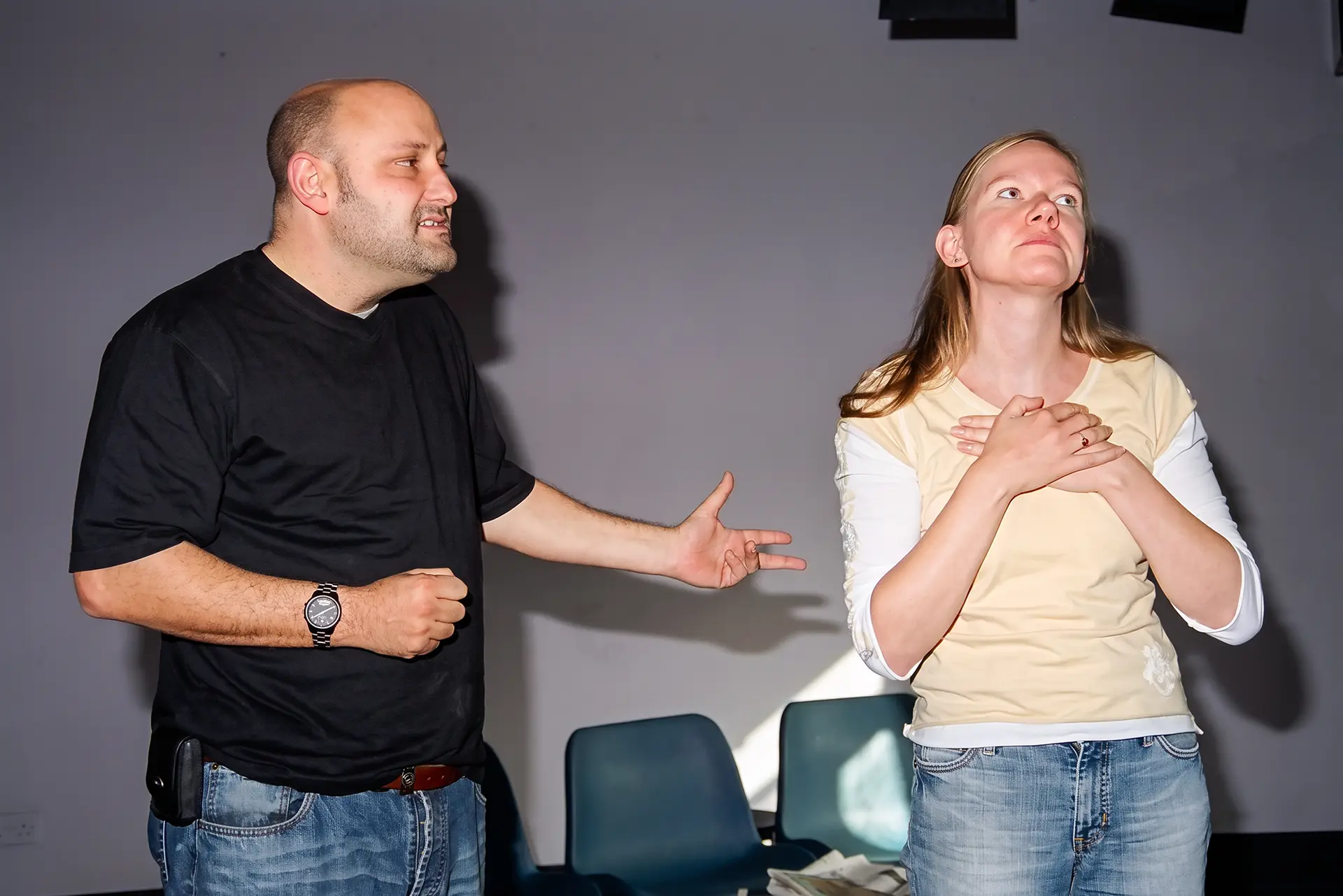 A man gestures with an open palm towards a woman standing with both hands pressed to her chest, looking up and away, rehearsing the cheesy curtain call episode at the Alma Tavern.
