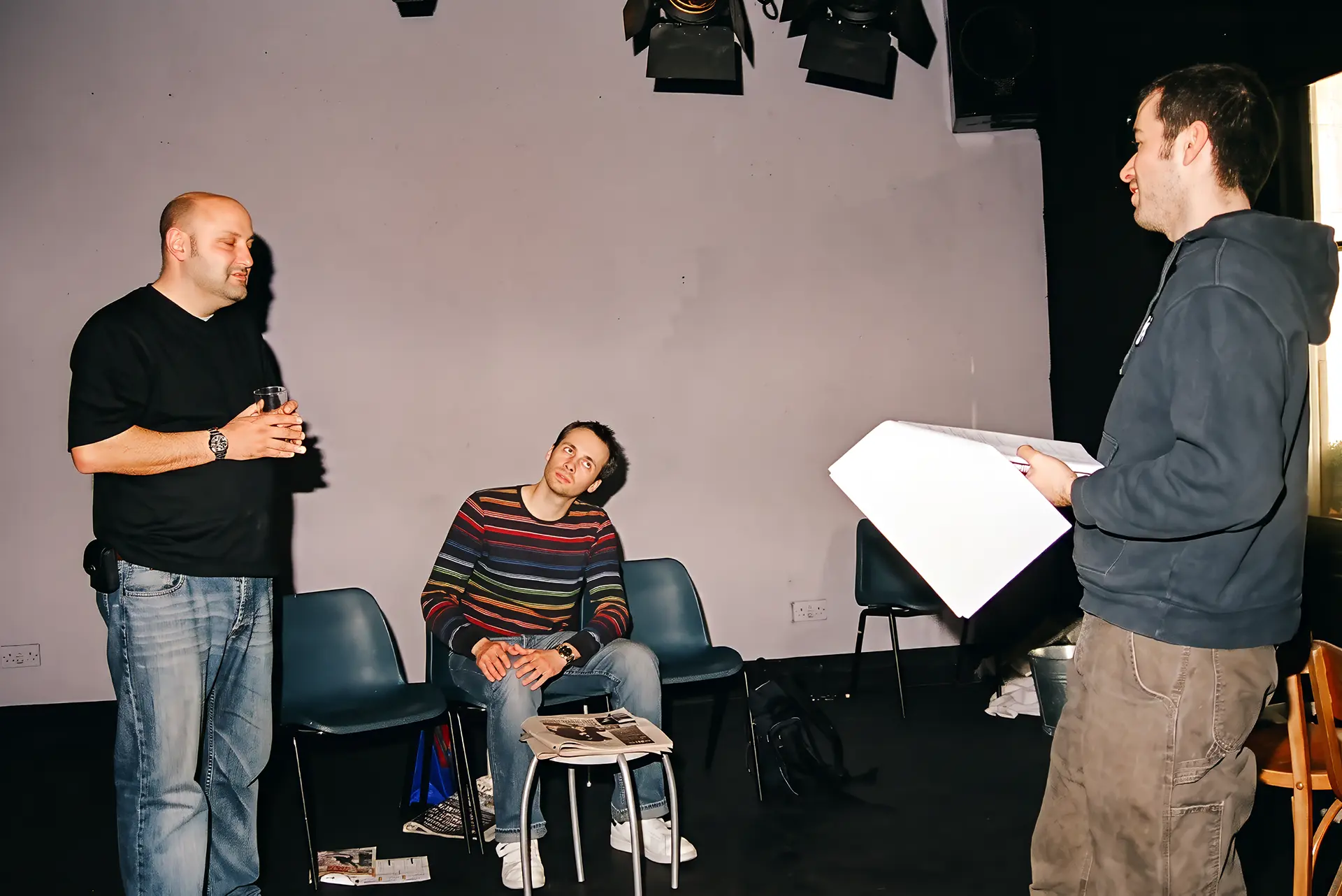 The director stands at the right of the frame holding an open script, speaking to two actors on the Alma Tavern stage with lighting lanterns rigged overhead: one stands at the left holding a pint glass in both hands and looking down; the other sits on a stacking chair, head inclined towards the standing actor but his eyes on the director.