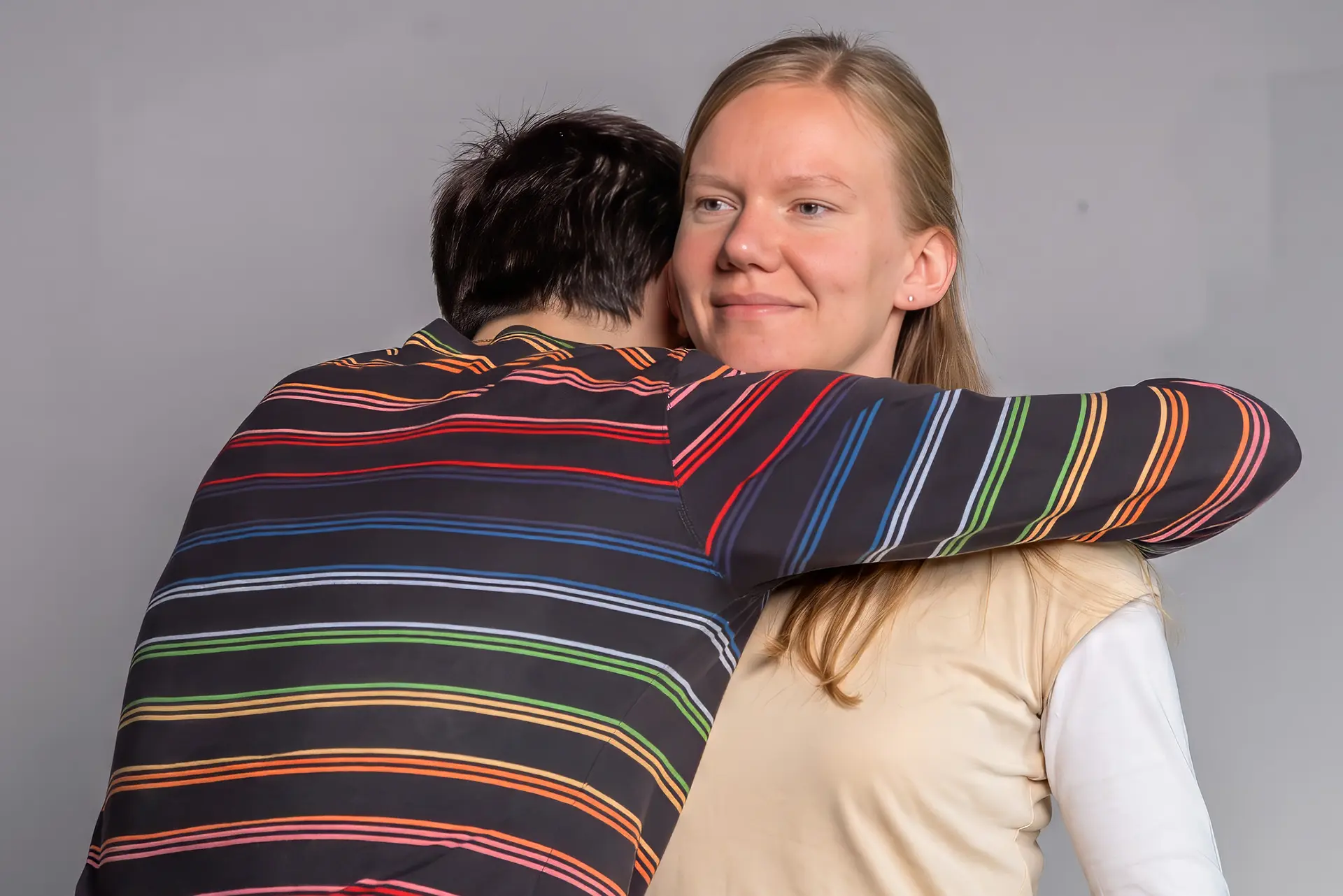 A man embraces a woman with both arms, his back to the camera; she leans back out of the embrace without returning it, looking away with a slight, unreadable smile.