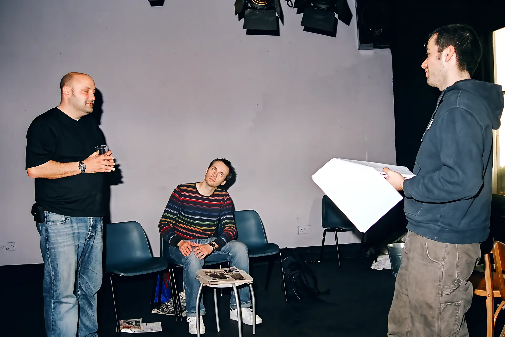 The director stands at the right of the frame holding an open script, speaking to two actors on the Alma Tavern stage with lighting lanterns rigged overhead: one stands at the left holding a pint glass in both hands and looking down; the other sits on a stacking chair, head inclined towards the standing actor but his eyes on the director.