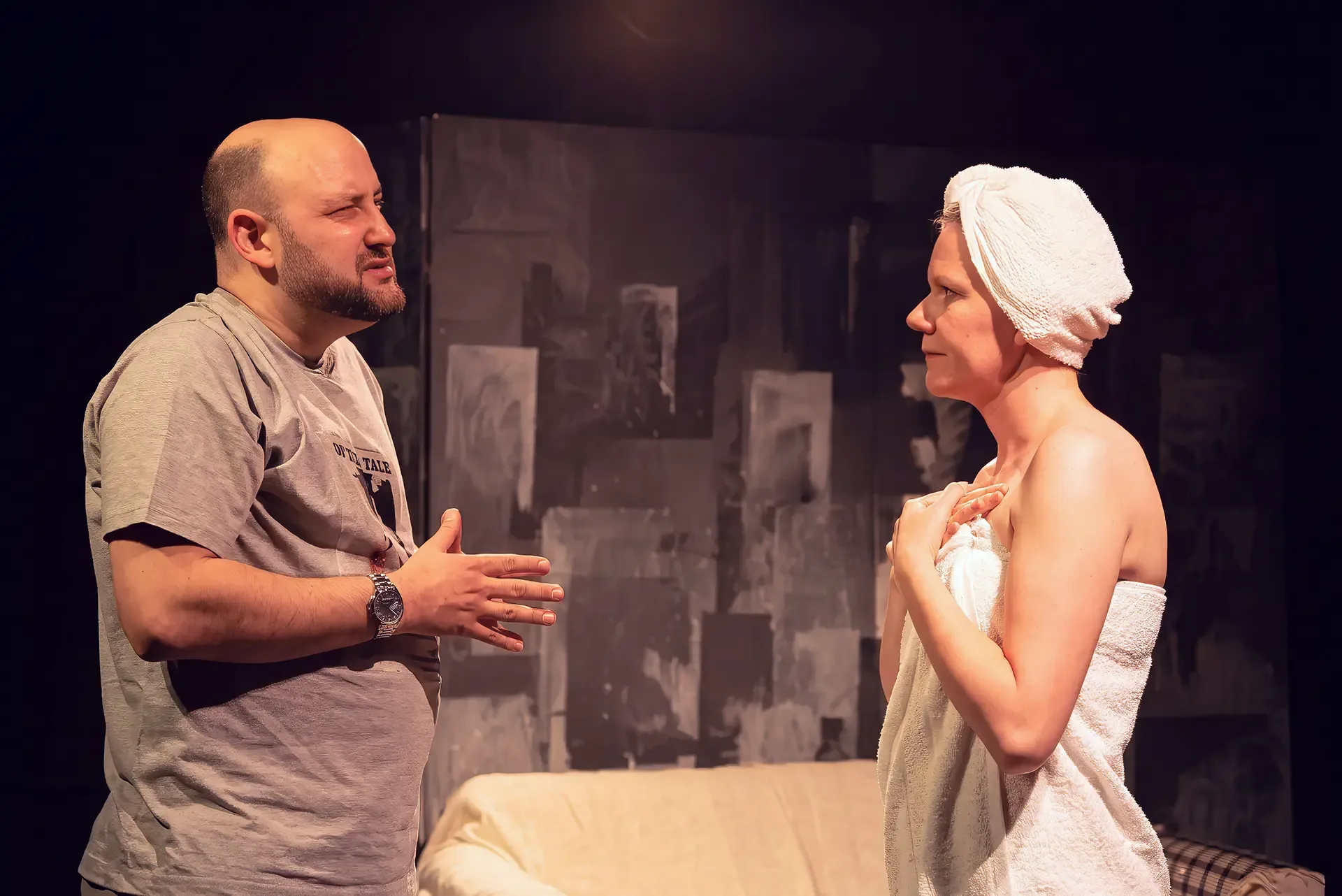 A man and a woman face each other on stage in front of grey block-patterned flats. The man, on the left, wears a grey printed T-shirt and a metal wristwatch; both hands are raised in front of his chest, fingertips touching. The woman on the right is wrapped in a white bath towel, with a second towel twisted around her hair, holding the towel closed at her chest with both hands. A sofa with a cream cover stands behind them.