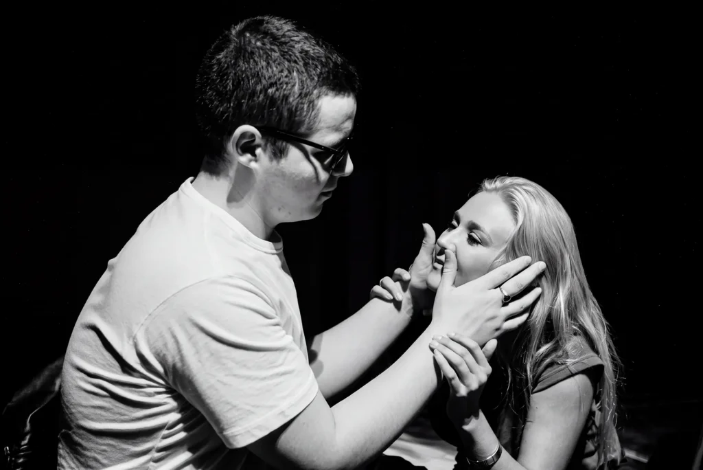 A black-and-white photograph of a young man in a white T-shirt and dark glasses cupping a young woman's face with both hands as she reaches up to touch his fingers